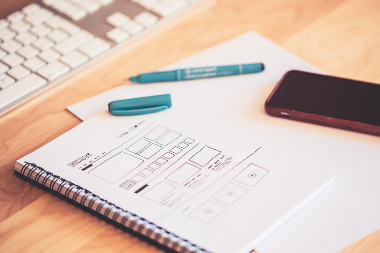 Notebook sketch of a website wireframe with uncapped pen, blank sheets of paper and cell phone on a desk.