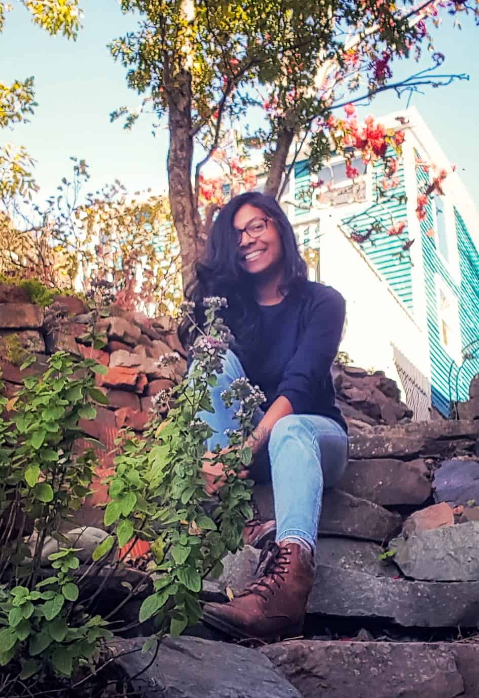 Ruha is wearing a navy sweater, jeans and brown boots, sitting on worn stone steps in a fall garden
